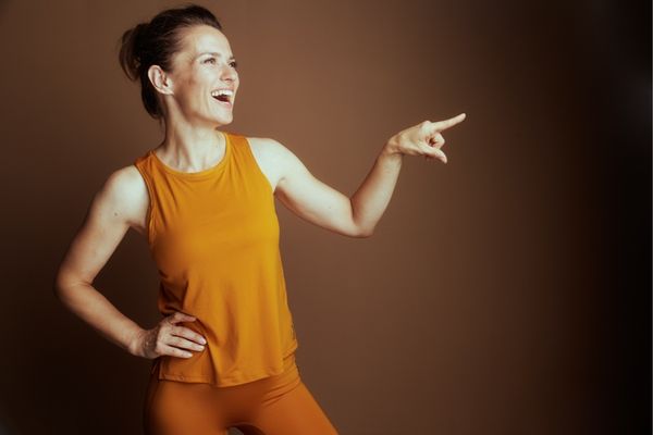 Woman in orange tank top and pants pointing to the side against a brown background